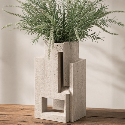 PLIOXOO® travertine geometric sculptural vase on wooden table with dried greenery – full product view showing stepped rectangular planes and narrow square opening in warm cream stone